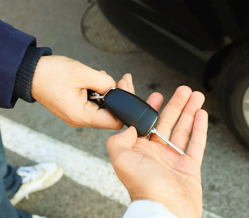 Person handing over a car key to another person outdoors near a vehicle tire and road markings.